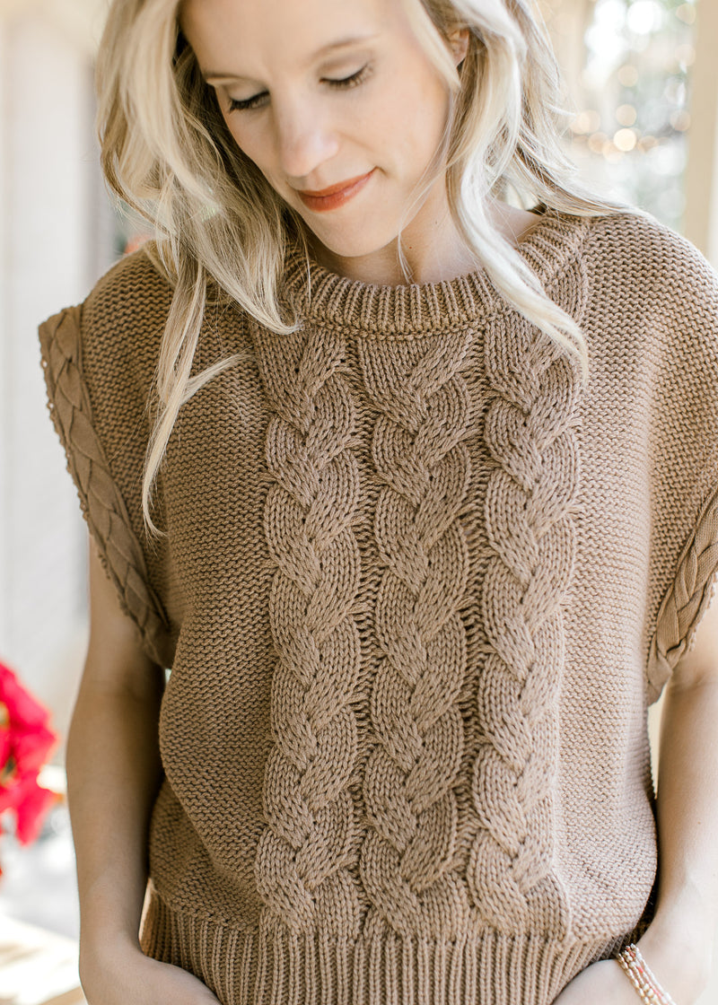 Model wearing a mocha cable knit sweater with capped sleeves and a round neck. 