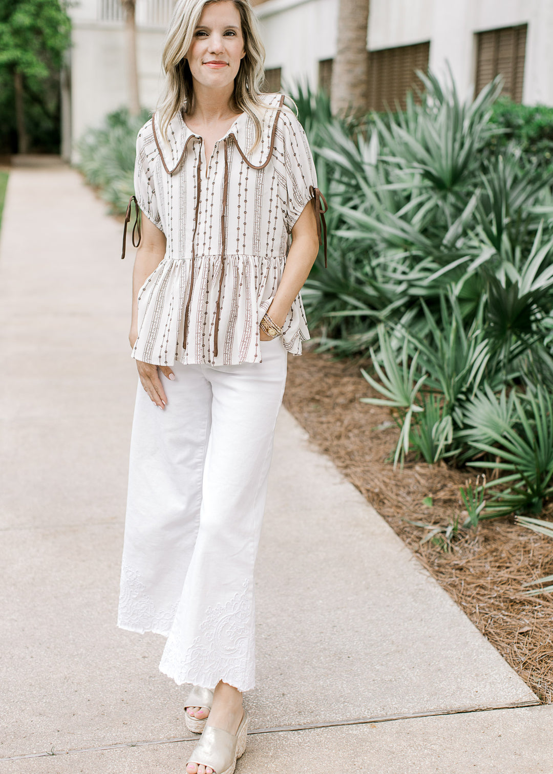 Model wearing a taupe top with brown floral, flap collar with tie and short sleeves with tie.