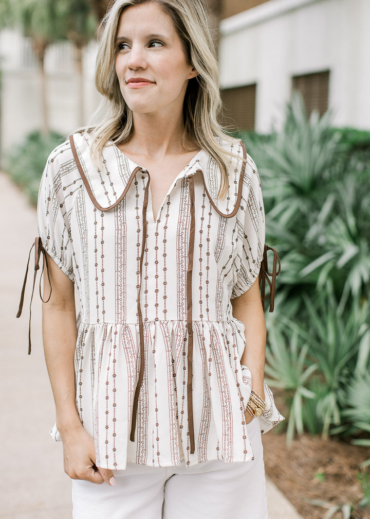 Model wearing a cream babydoll blouse with brown floral, flap collar, v-neck and short sleeves.