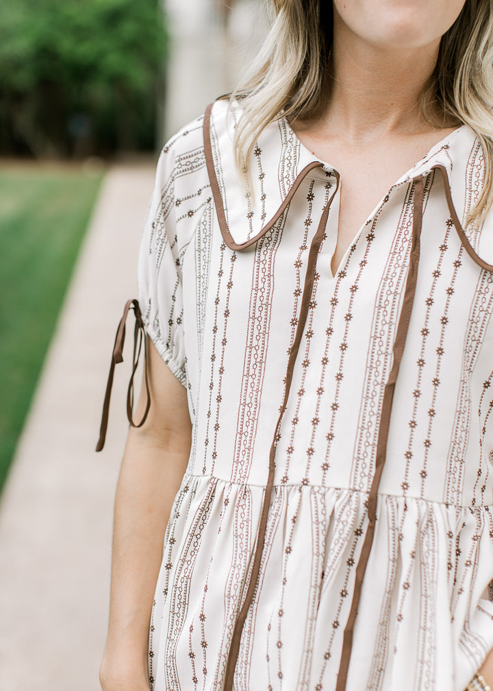 Close up of v-neck and flap collar with tie on a cream blouse with brown floral pattern. 