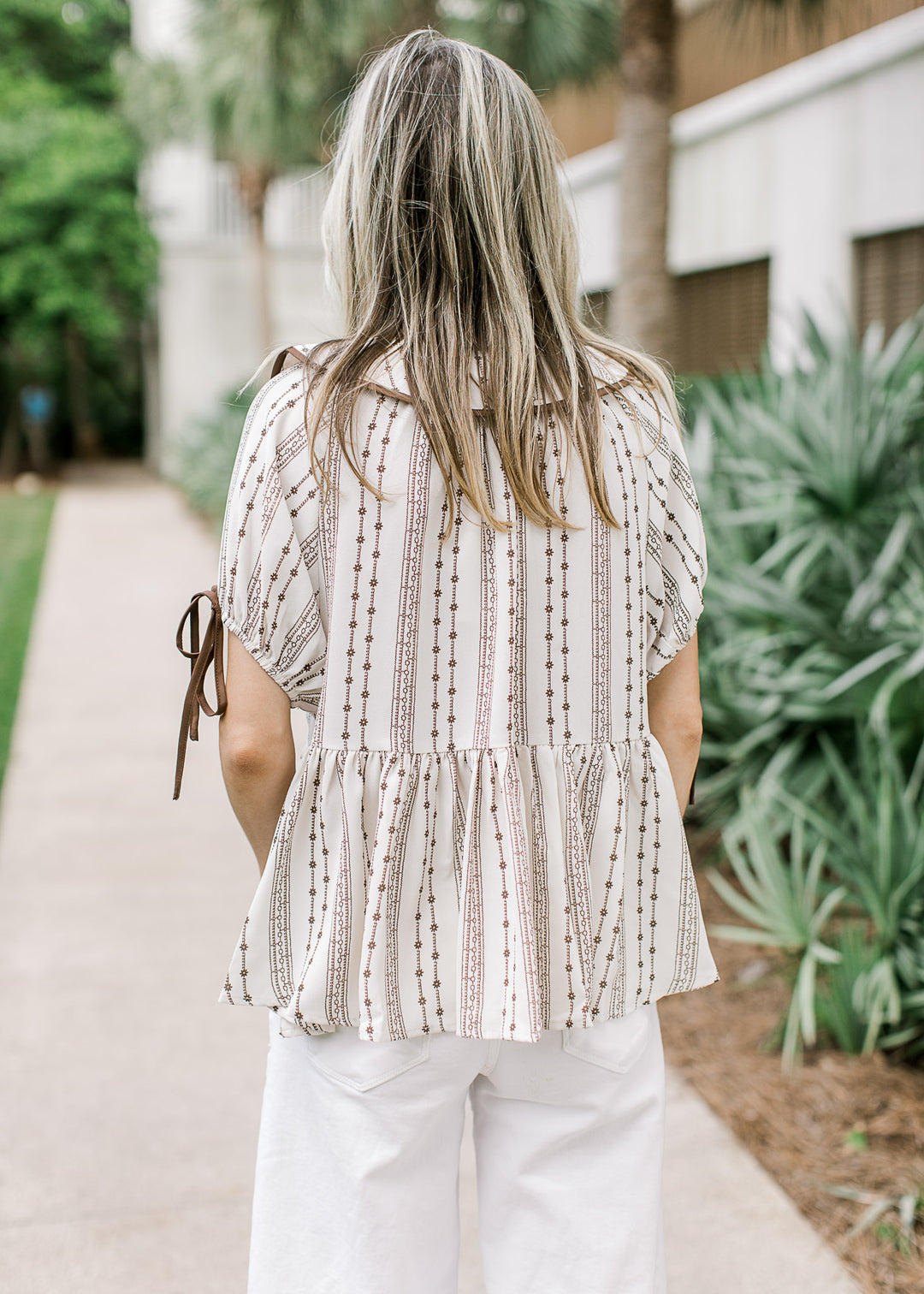 Back view of Model wearing a taupe top with brown floral, flap collar and short sleeves with tie.