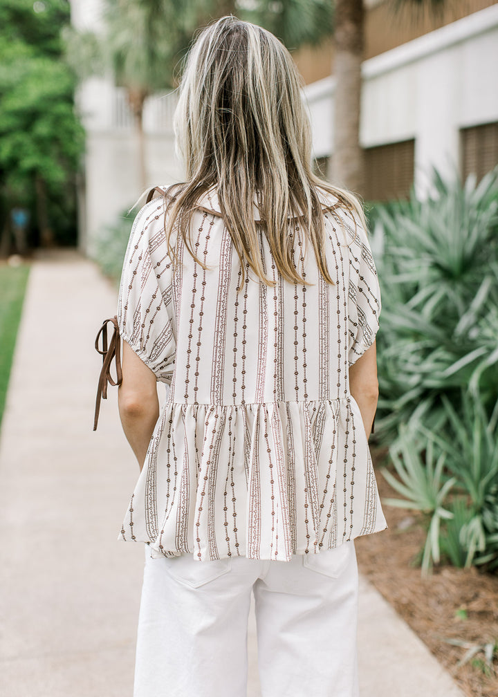 Back view of Model wearing a taupe top with brown floral, flap collar and short sleeves with tie.