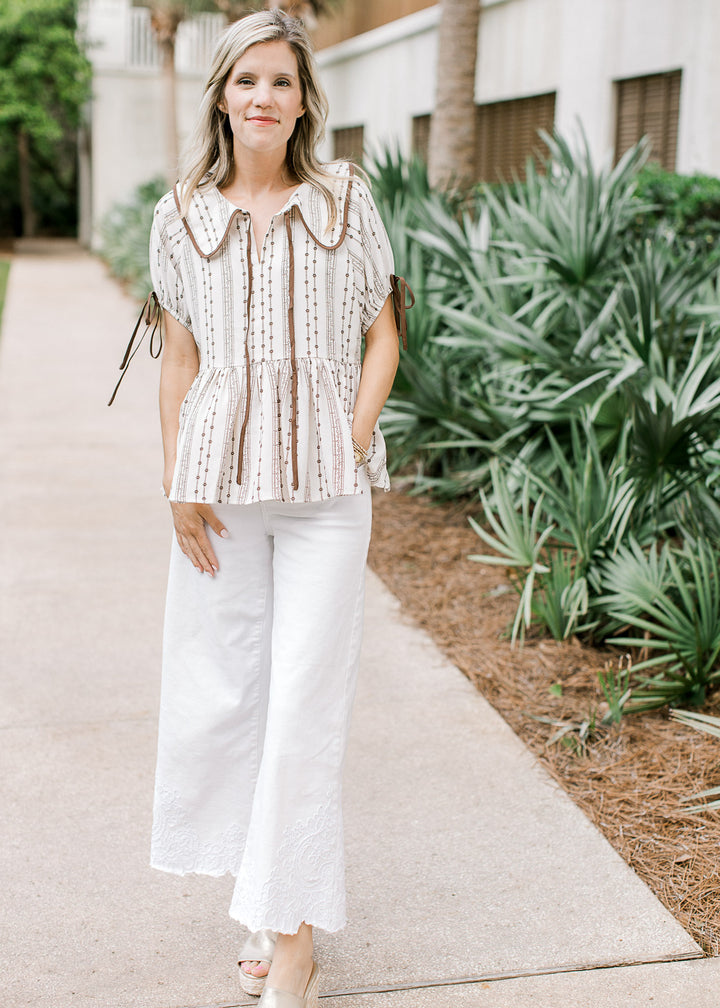 Model wearing white pants and a taupe top with brown floral, collar and short sleeves with tie.