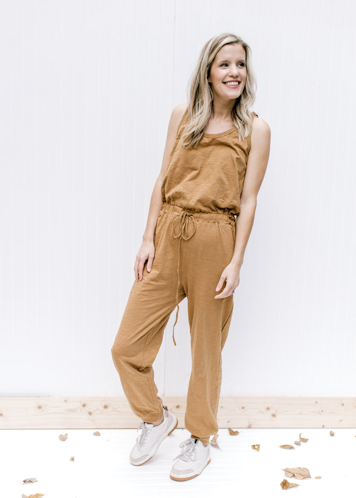 Model wearing a sleeveless mocha jumpsuit with a drawstring waist, pockets and elastic at leg.
