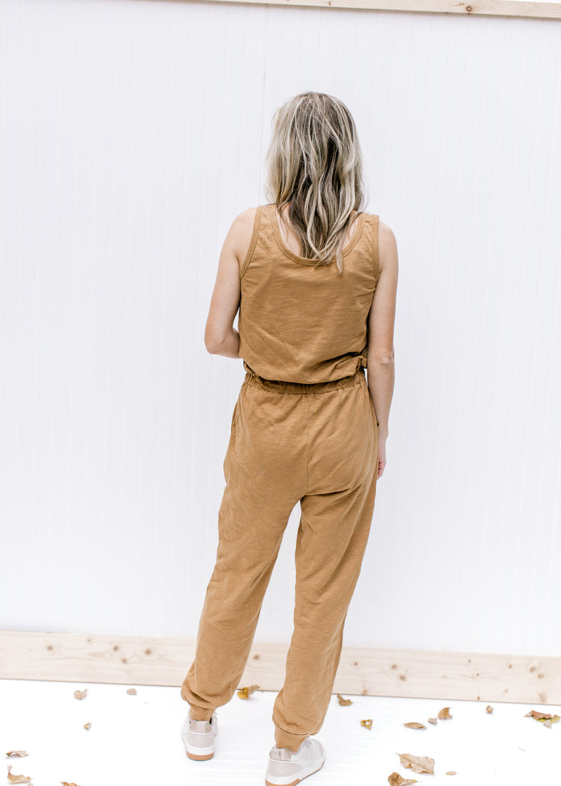 Back view of Model wearing a sleeveless mocha jumpsuit with pockets and elastic at leg.