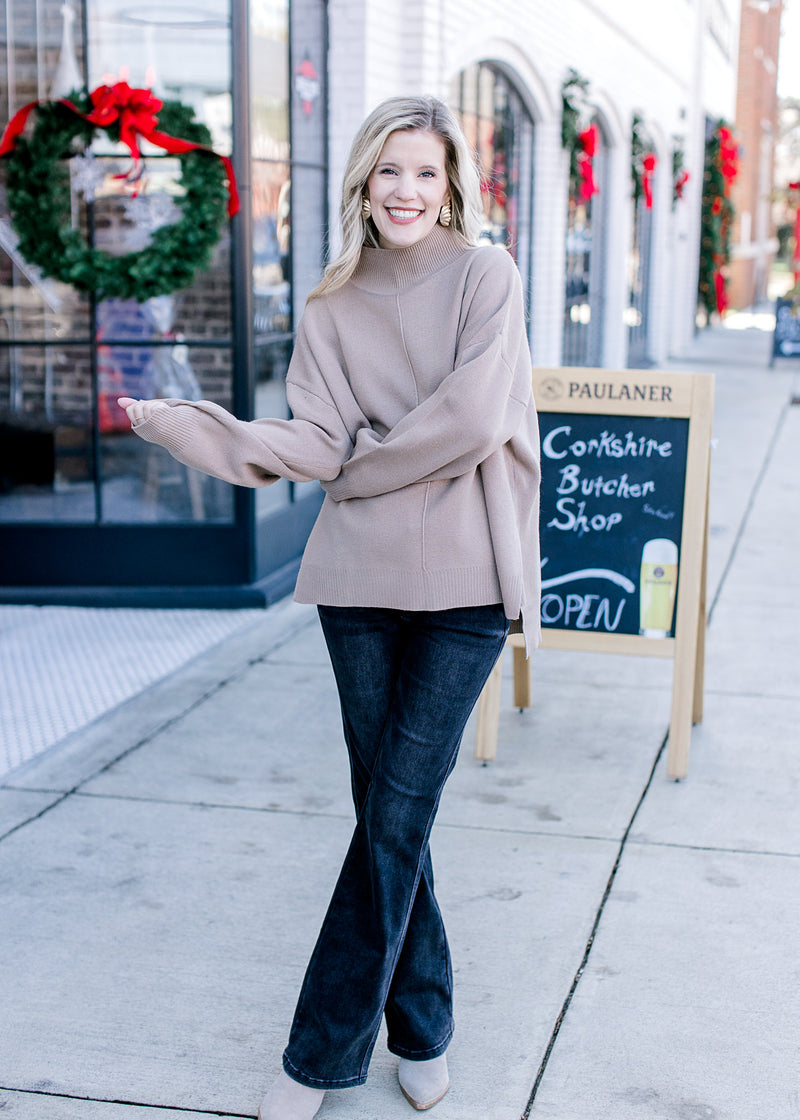 Model wearing jeans and a mocha sweater with a mock neck, long sleeves and extended shoulder. 