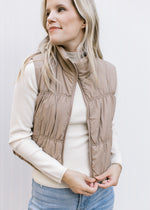 Model wearing a mocha puffer vest with a zipper, pockets and a mock neck.