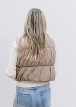 Back view of Model wearing a mocha puffer vest with a zipper, pockets and a mock neck.