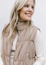 Close up of Model wearing a mocha puffer vest with a zipper, pockets and a mock neck.