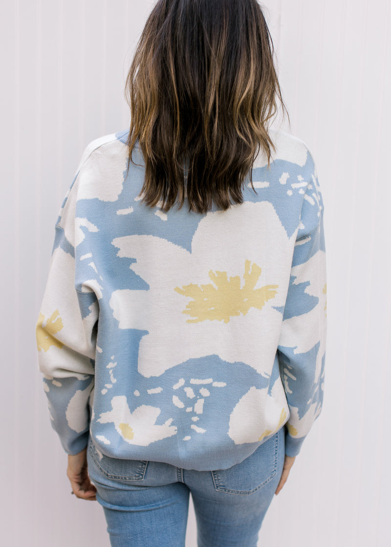 Back view of Model wearing a pale blue sweater with cream and yellow floral and long sleeves. 