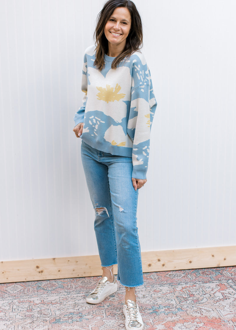 Model wearing jeans, sneakers and a pale blue long sleeve sweater with cream and yellow floral.