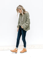 Model wearing jeans and booties with a moss hoodie with a woven white string detail at the hood. 