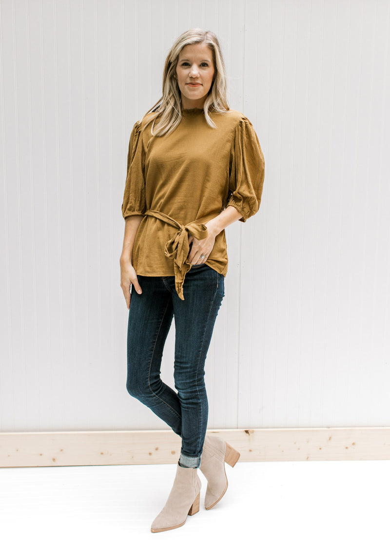 Model wearing jeans, booties and a copper colored short sleeve top with a ruffled neck and a tie. 
