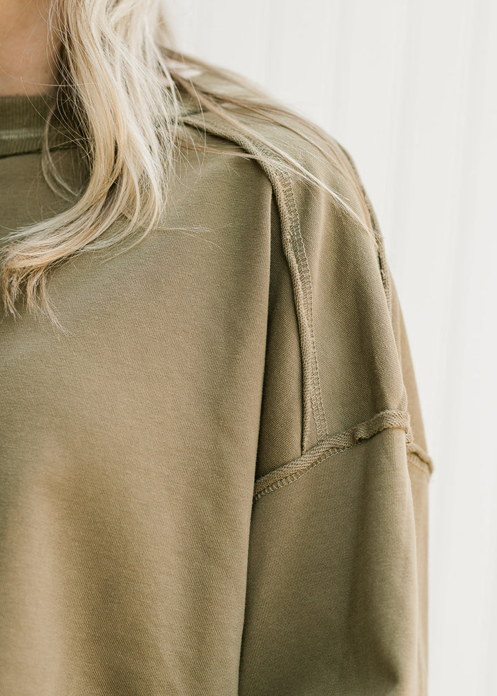 Close up of exposed hem and extended shoulder on an olive lightweight top with a round neck. 