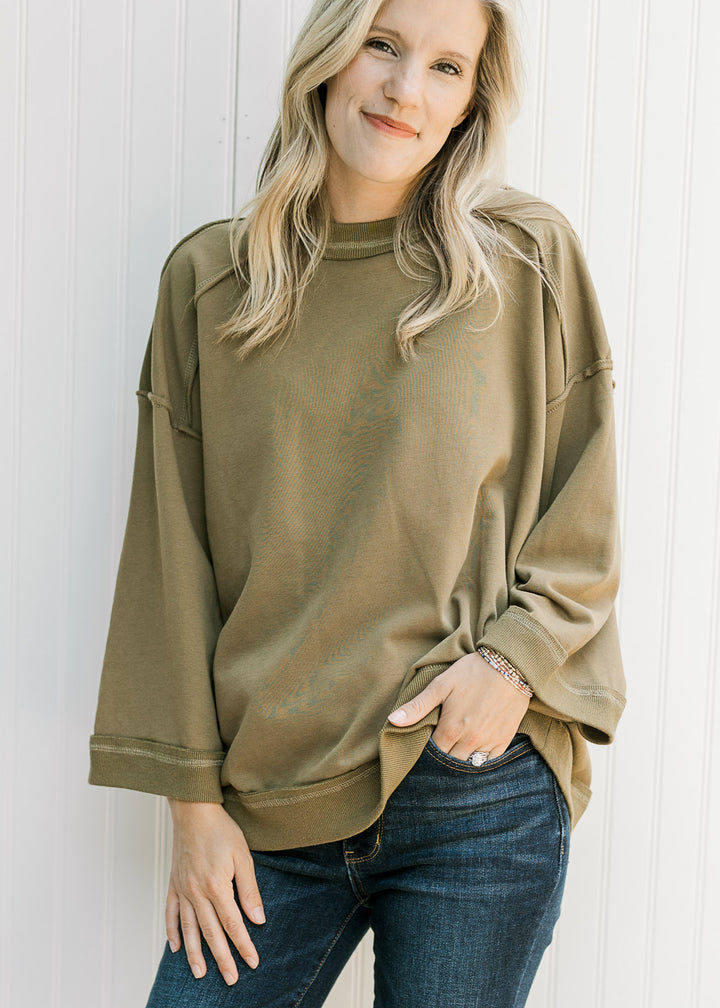 Model wearing jeans and an olive 3/4 sleeve top with exposed hem and patch with seam along back.