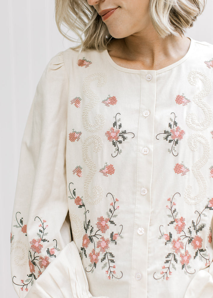 Close up of rose, taupe, and green cross-stitch embroidery on a taupe button up long sleeve top.