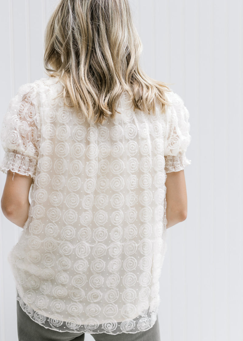 Back view of Model wearing a cream v-neck top with floral lace overlay and lace short sleeves.