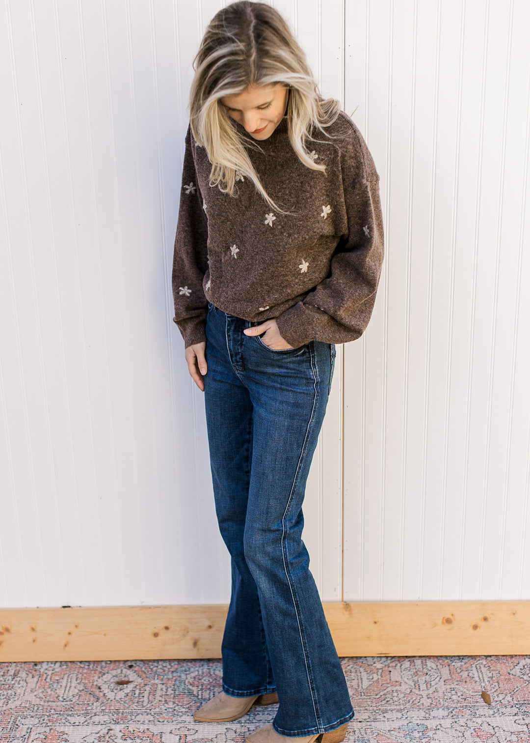 Model wearing jeans and a brown sweater with taupe stitched flowers, a mock neck and long sleeves. 