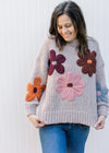 Model wearing a taupe sweater with multicolored yarn flowers, long sleeves and round neck.