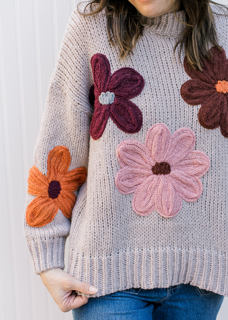 Close  up of multi colored yarn flowers on a taupe knit sweater with long sleeves.