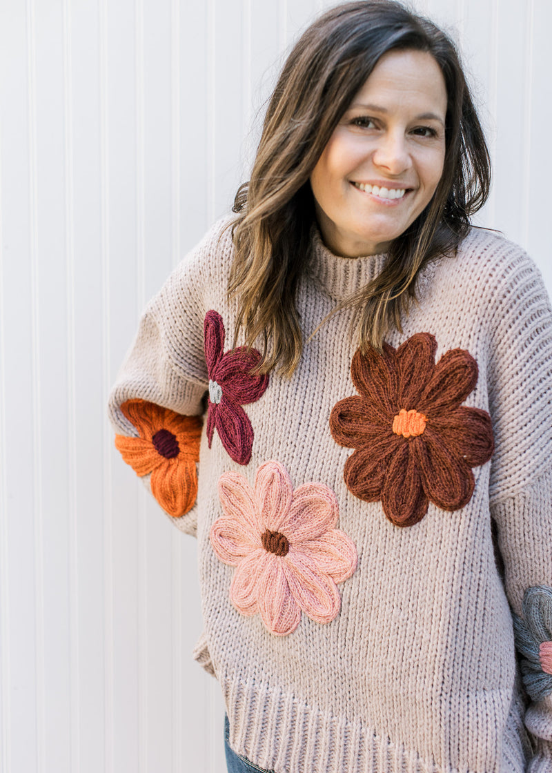 Model wearing a taupe knit sweater with multicolored yarn flowers, long sleeves and round neck.