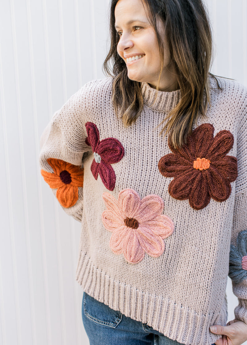 Model wearing jeans and a taupe sweater with multicolored yarn flowers, long sleeves and round neck.