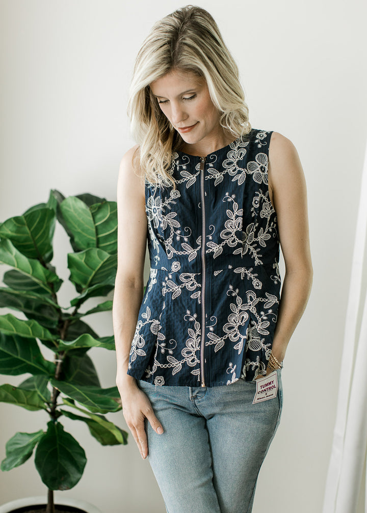  Model wearing a navy sleeveless top with embroidered floral, front zipper and pleating at bust.