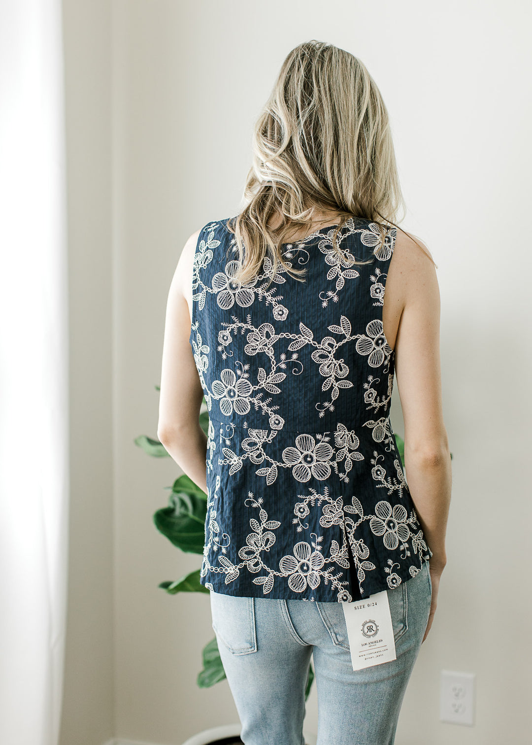 Back view of Model wearing a navy sleeveless top with embroidered floral, pleated hem.