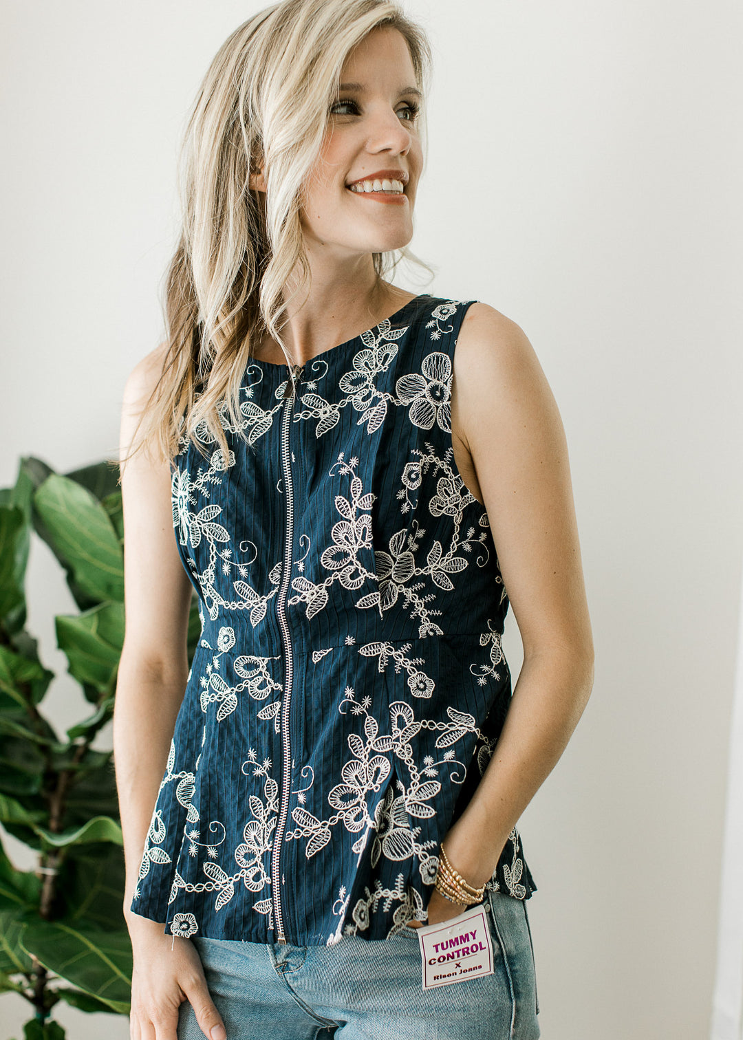 Model wearing jeans and a navy sleeveless top with floral, front zipper and pleating detail.