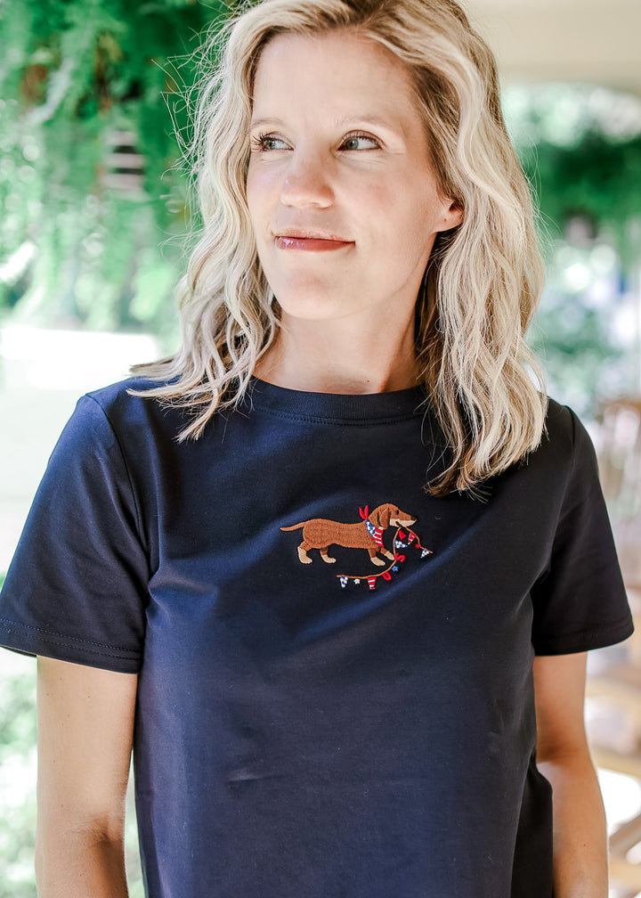 Model wearing a navy short sleeve top with an embroidered Dachshund with USA banner. 