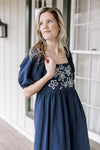 Model wearing a nay dress with a square neck, embroidered flowers and bubble short sleeves.