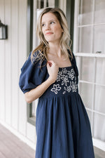 Model wearing a nay dress with a square neck, embroidered flowers and bubble short sleeves.