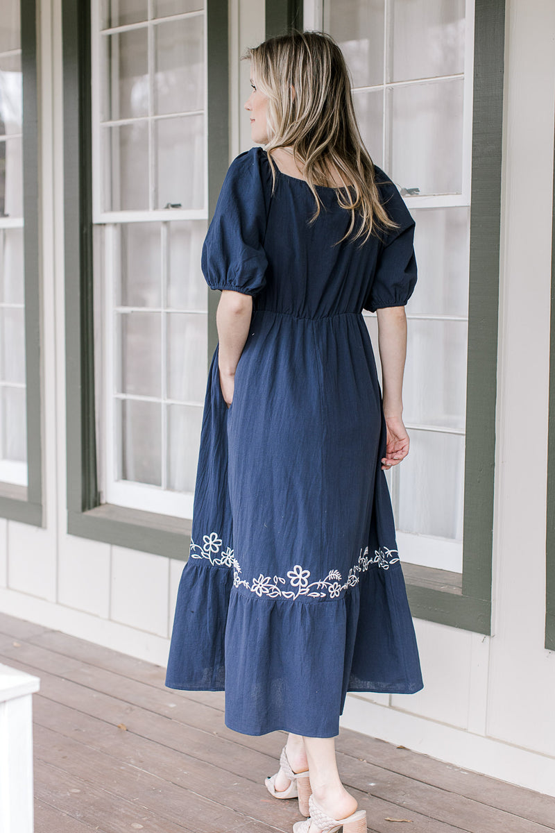 Back view of a navy midi with a square neck, bubble short sleeves and white embroidered detail. 