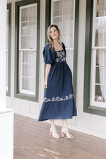 Model wearing heels and a navy dress with a square neck, embroidered flowers and short sleeves.