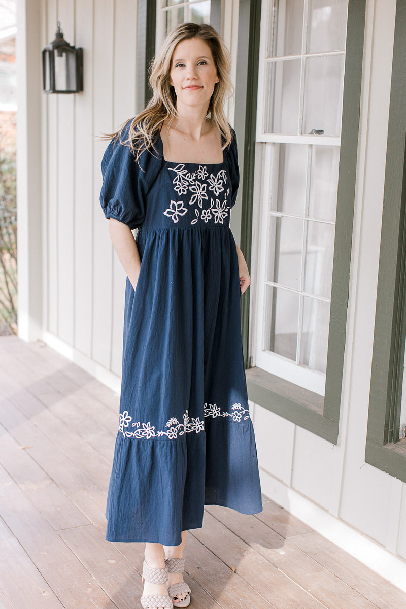 Model wearing a navy dress with a square neck, embroidered white flowers and pockets. 