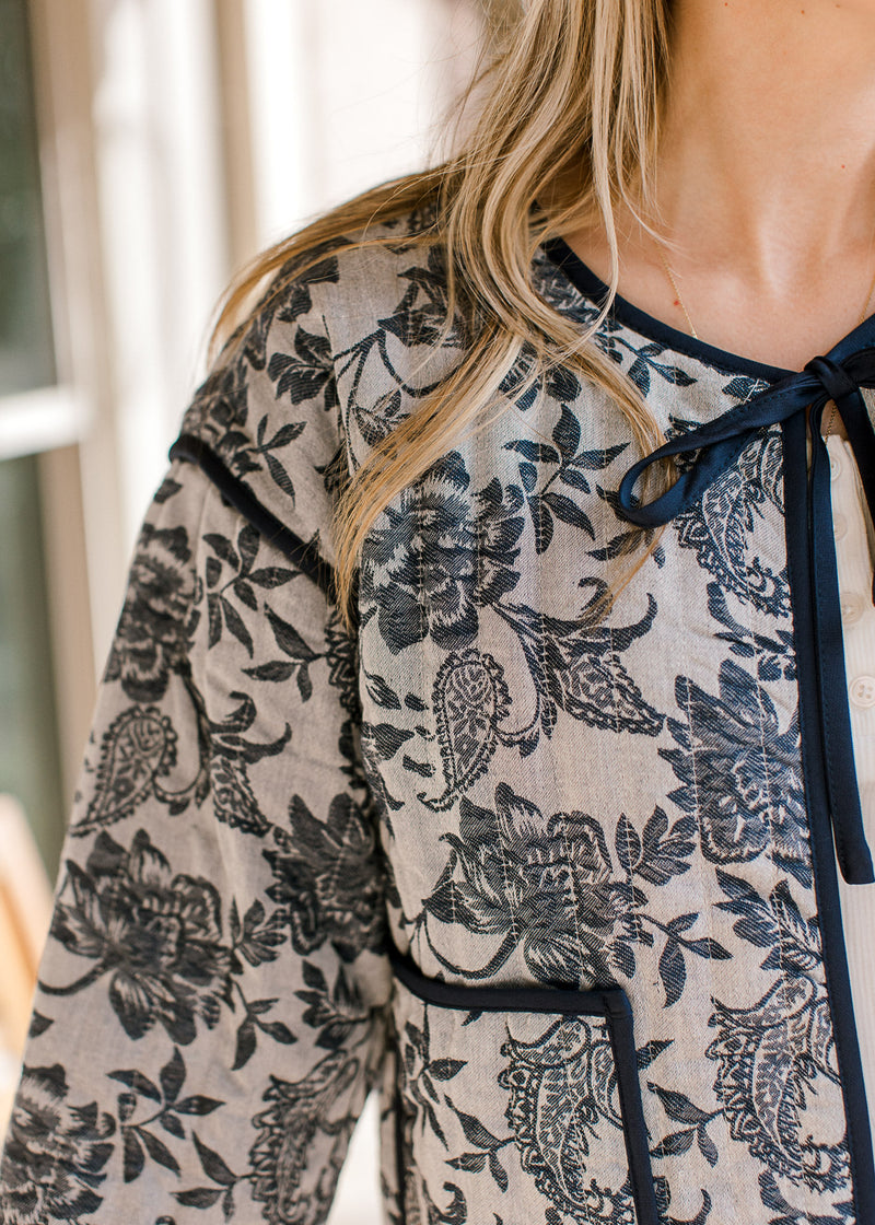 Close up of tie closure and quilted fabric on a gray jacket with navy floral and long sleeves. 