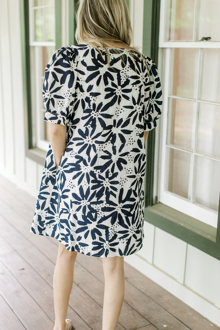 Back view of Model wearing an off white dress with navy flowers, short puff sleeves and a v-neck.