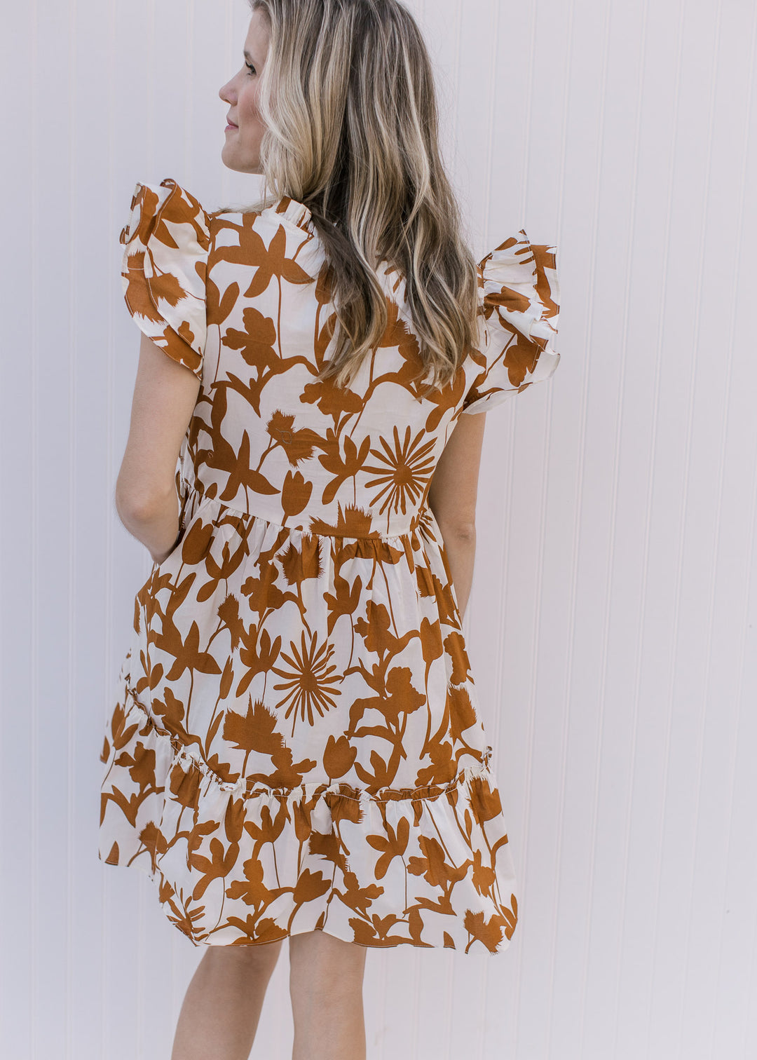 Back view of Model wearing a cream dress with rust floral pattern, ruffle short sleeves,and pockets.