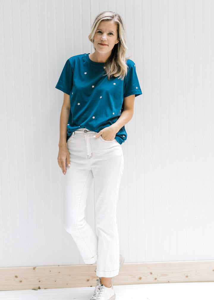 Model wearing white jeans with sneakers and a navy top with cream embroidered stars. 