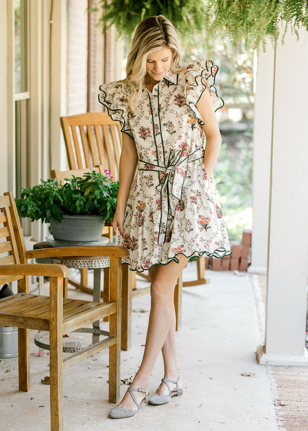 Model wearing heels and a cream dress with floral print, button front and ruffle cap sleeves.