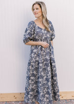 Model wearing a gray, v-neck maxi with navy floral, short puff sleeves and pockets. 