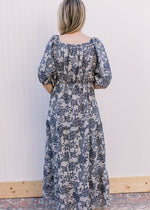 Back view of Model wearing a gray maxi with navy floral, short puff sleeves and an elastic band.