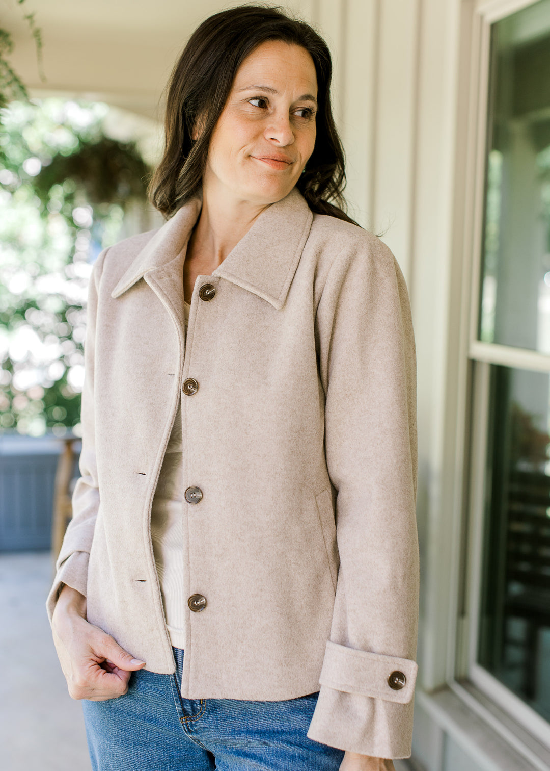 Model wearing a long sleeve oatmeal jacket with brown buttons, flap collar and side slit pockets.