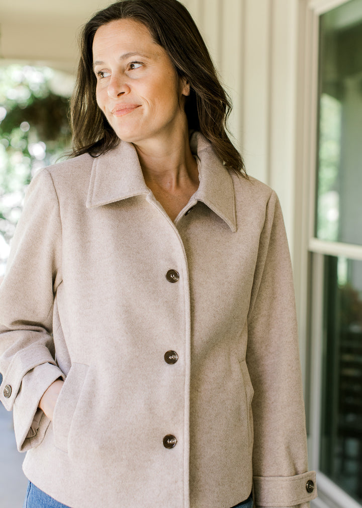 Model wearing a buttoned up long sleeve oatmeal jacket with a flap collar and side slit pockets.