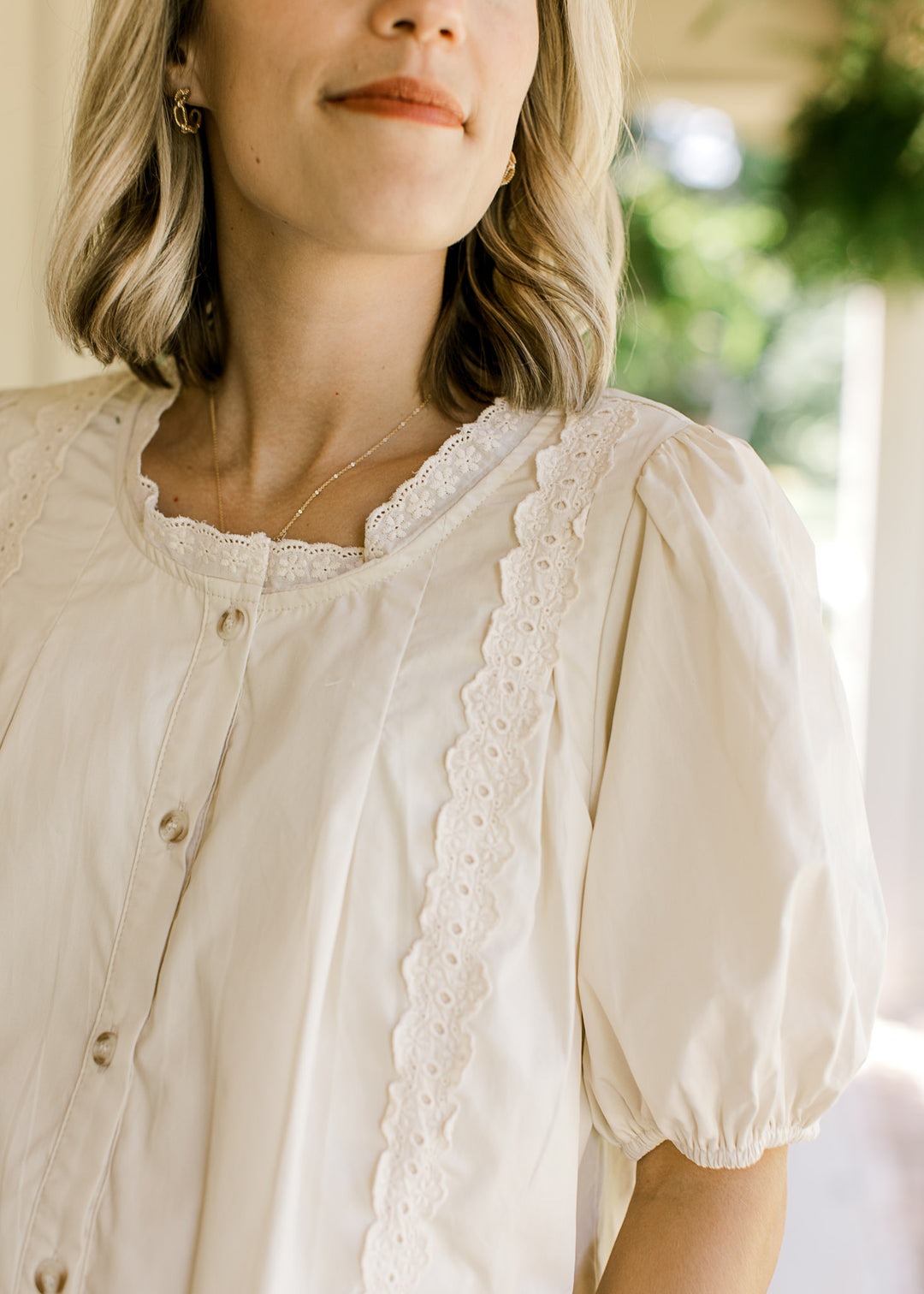Close up of lace at round neck and outer front on an oatmeal button up top with short sleeves.