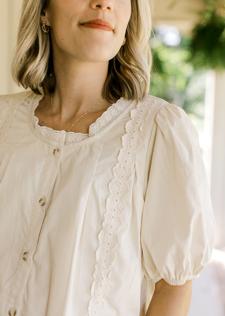 Close up of lace at round neck and outer front on an oatmeal button up top with short sleeves.