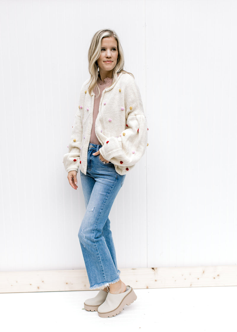 Model wearing an oatmeal open front cardigan with bubble long sleeves and colored pop-poms. 