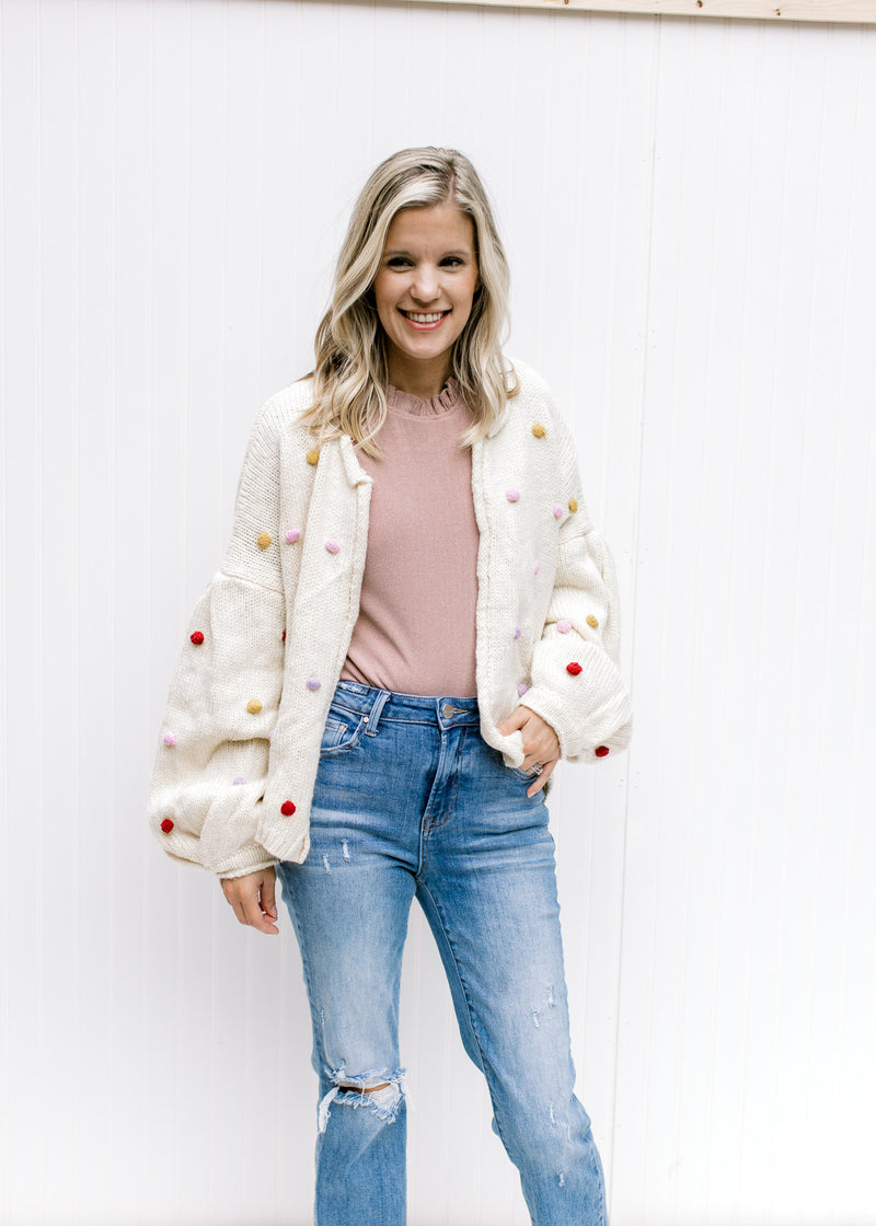 Model wearing a blush top with an oatmeal cardigan with bubble long sleeves and colored pop-poms. 