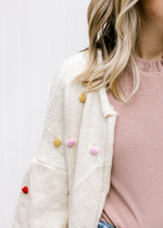 Close up of mustard, pink, lavender and maroon Pom-poms on an oatmeal open front cardigan. 