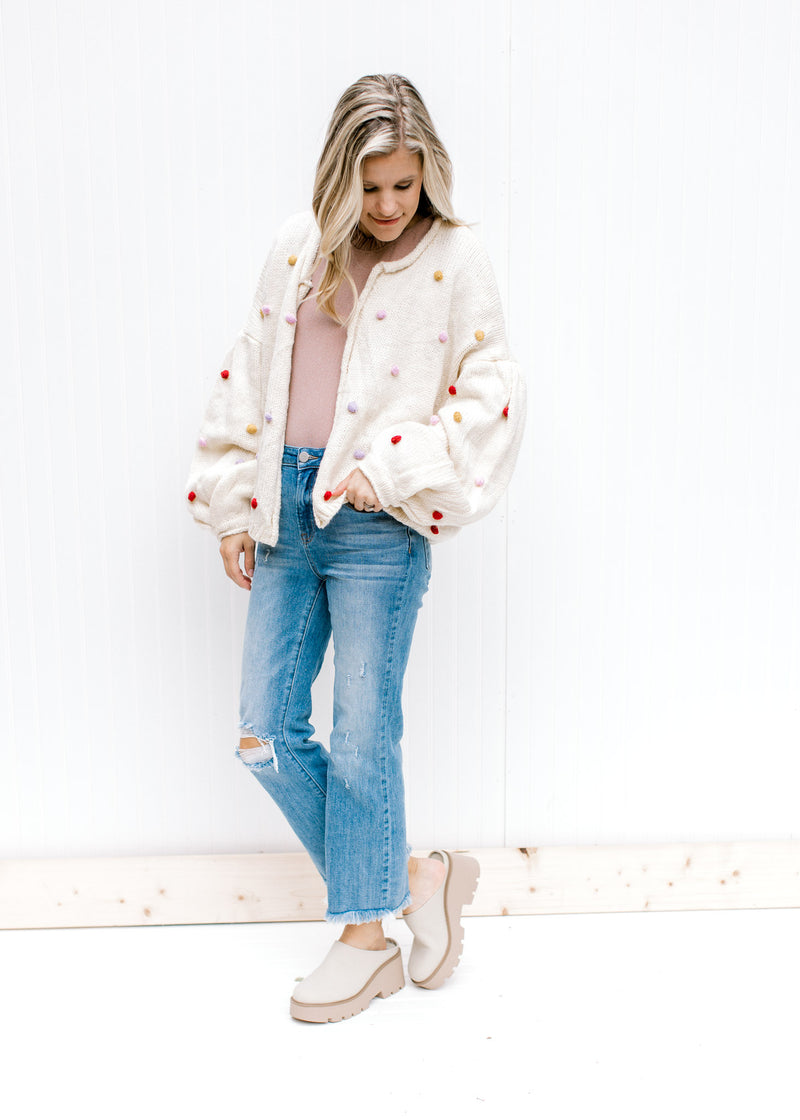 Model wearing jeans and mules with an oatmeal cardigan with long sleeves and colored pop-poms. 